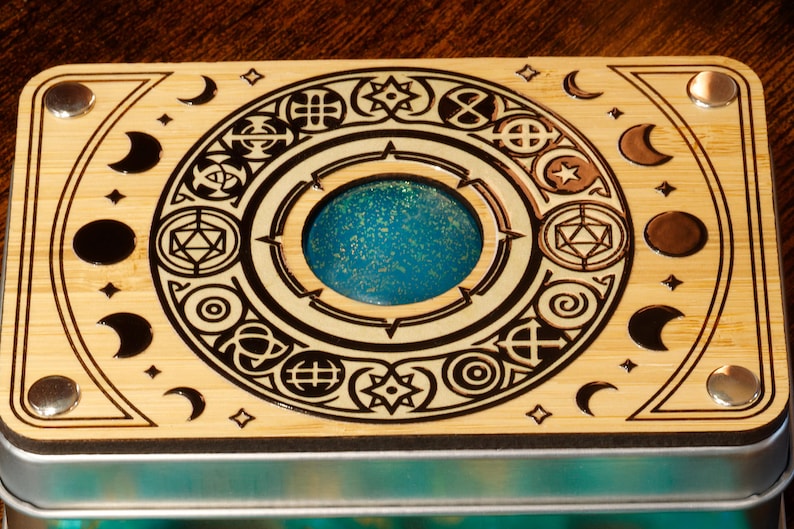 K&ouml;nnte beinhalten: Eine Holzkiste mit einem kreisf&ouml;rmigen Design, das verschiedene Symbole und eine blaue, glitzernde Mitte zeigt. Die Kiste hat einen Metallrahmen und vier Metallakzente.