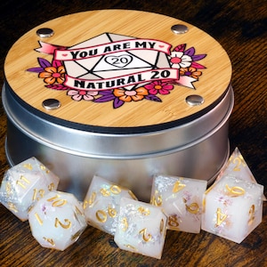 May include: A round, silver tin with a wooden lid featuring the text "You Are My Natural 20" surrounded by floral and banner designs. Several clear, glittery dice with gold numbers are in front of the tin.