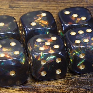 Könnte beinhalten: Ein Set aus fünf durchscheinenden schwarzen Würfeln mit abgerundeten Ecken und goldfarbenen Augen. Das Innere jedes Würfels enthält irisierende, reflektierende Folienflocken, die bei Lichteinfall in Grün-, Blau- und Orangetönen schimmern.