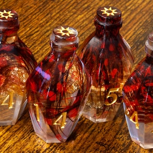Könnte beinhalten: Vier dekorative Glasflaschen mit roten und goldenen federartigen Einschlüssen. Jede Flasche hat ein goldenes Sternmuster auf dem Deckel und eine goldene Zahl auf der Basis. Die Flaschen stehen auf einer Holzoberfläche.