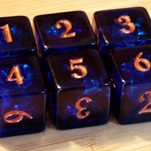 Pode incluir: Um conjunto de seis dados de seis faces azul-escuro translúcidos dispostos em duas filas. Cada dado apresenta números gravados preenchidos com um pigmento de cor cobre metálico. Os números de um a seis são visíveis nas faces.
