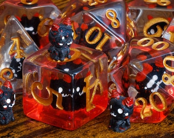 May include: A set of polyhedral dice made of clear resin with a red base, featuring embedded black and red horned creature figurines. The dice have gold-colored numbers and include a d20, d12, d10, d8, d6, and d4, with small creature figures placed around them.