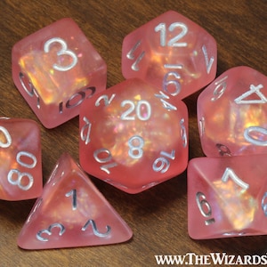 May include: A set of seven translucent pink polyhedral dice with iridescent internal flecks and silver-colored numbers. The dice include a d20, d12, d10, d8, d6, d4, and a percentile die. The text WWW.THEWIZARDSVAULT.COM is printed in the bottom right corner.