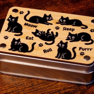 以下が含まれることがあります： 「Meow」「Sleep」「Eat」「Roll」「Purrr」などの言葉と、さまざまな行動をとる黒い猫のシルエットが描かれた、金属製の蓋が付いた木製の箱。