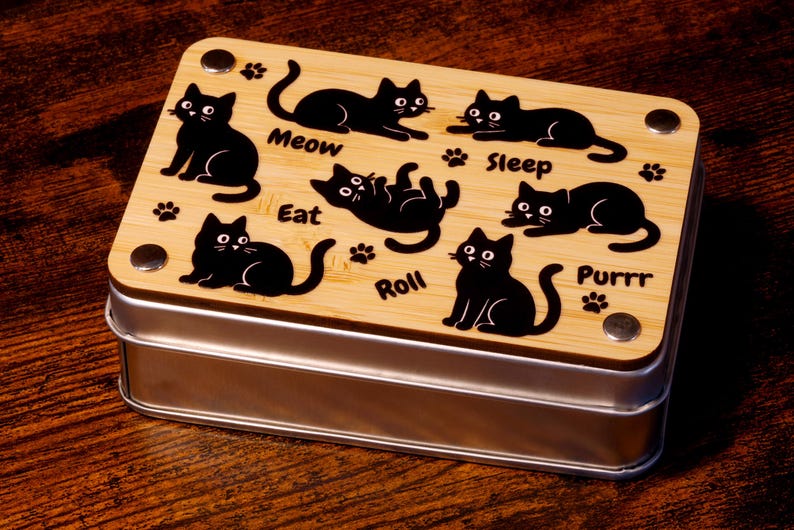 以下が含まれることがあります： 「Meow」「Sleep」「Eat」「Roll」「Purrr」などの言葉と、さまざまな行動をとる黒い猫のシルエットが描かれた、金属製の蓋が付いた木製の箱。
