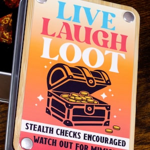 May include: A metal dice box with a wooden lid. The lid has a colorful graphic with the text "LIVE LAUGH LOOT" and an illustration of a treasure chest with gold coins. The text below the illustration reads "STEALTH CHECKS ENCOURAGED WATCH OUT FOR MIMICS".