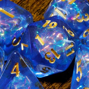 Op de afbeelding: Een verzameling doorschijnende blauwe veelzijdige dobbelstenen met een iriserend, glinsterend effect aan de binnenkant en gegraveerde goudkleurige cijfers. De set bevat diverse geometrische vormen met scherpe randen en een reflecterende uitstraling.