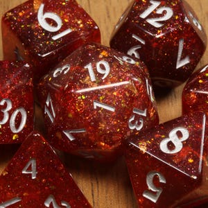 Peut inclure: Un ensemble de sept dés polyédriques en résine rouge foncé translucide avec des paillettes dorées incrustées. Chaque dé présente des chiffres gravés en blanc, dont un d20 affichant le nombre 19 et un d100 montrant clairement les nombres 70 et 30.
