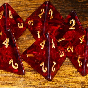 May include: Six red, translucent, four-sided dice with gold-colored numbers. The dice are arranged on a wooden surface, showcasing their geometric shape and color contrast. Ideal for tabletop games.