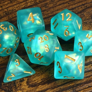 May include: A set of seven polyhedral dice in a translucent teal colour with an iridescent finish. Each die features engraved numbers filled with gold-coloured paint, including a d20, d12, d10, d8, d6, d4, and a percentile die with tens digits.