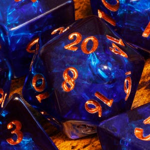 May include: Close-up of several polyhedral dice. The dice are a deep blue with orange numbering. The dice have a marbled appearance, with swirls of lighter blue and white. The dice are arranged on a textured, golden-brown surface.