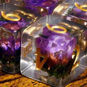 May include: A set of clear resin dice with rounded edges, each containing embedded purple flowers and green moss. The numbers on the faces are engraved and filled with a metallic gold pigment, featuring a distinct font style for each digit.