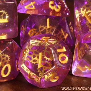 May include: A set of translucent purple polyhedral dice featuring embedded gold-colored metal gear mechanisms and gold-colored numbers. The text WWW.THEWIZARDSVAULT.COM is printed in the bottom right corner of the image.
