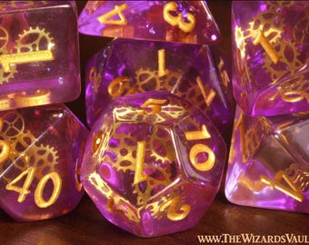 May include: A set of translucent purple polyhedral dice featuring embedded gold-colored metal gear mechanisms and gold-colored numbers. The text WWW.THEWIZARDSVAULT.COM is printed in the bottom right corner of the image.