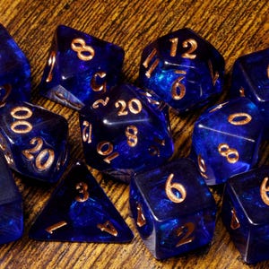 May include: A set of ten translucent deep blue polyhedral dice with gold-colored engraved numbers. The collection includes various shapes such as cubes, pyramids, and multi-faceted geometric forms, all featuring a smooth, glossy finish.