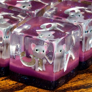 May include: A set of translucent purple resin dice featuring embedded miniature grey cat figurines. Each die has silver-colored numbers etched into the surface, with a dark purple glitter base layer visible at the bottom of each cube-shaped piece.