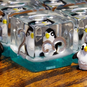 Op de afbeelding: Transparante harsblokjes met miniatuur zwart-witte pinguïnfiguurtjes met gele snavels. Elk blokje heeft een zilverkleurig cijfer aan de voorzijde en een doorschijnende blauwgroene basis. Twee losse pinguïnfiguurtjes staan voor de blokjes.