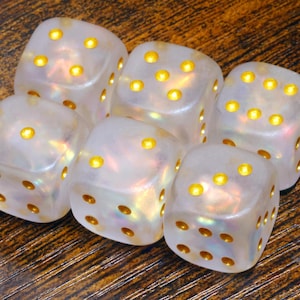 May include: A set of six translucent, pearlescent white dice with rounded corners. Each die features recessed circular pips filled with a bright, metallic gold colour. The material has a subtle iridescent sheen that reflects light across the surfaces.