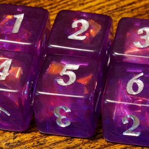 May include: A set of six translucent purple six-sided dice arranged in two rows of three. Each die features silver-coloured numerals engraved on the faces, with numbers one through six clearly visible across the top and front surfaces of the cubes.