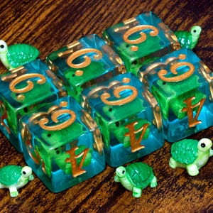 May include: A set of six translucent teal resin dice, each containing a small green turtle figurine inside. The dice feature gold-colored numbers engraved on the faces. Three additional small green turtle figurines are placed around the dice.