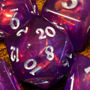 Può includere: Un set di dadi poliedrici viola traslucidi con texture interne scintillanti e numeri color argento. Il dado centrale a venti facce mostra il numero venti, circondato da altri dadi che presentano vari valori numerici incisi.