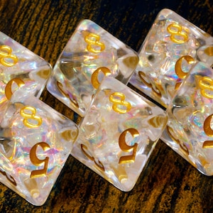 May include: Six clear, iridescent dice with gold-coloured numbers, mainly displaying the numbers 5 and 8. The dice are diamond-shaped and have a textured, crystalline appearance. The background is a dark, wooden surface.