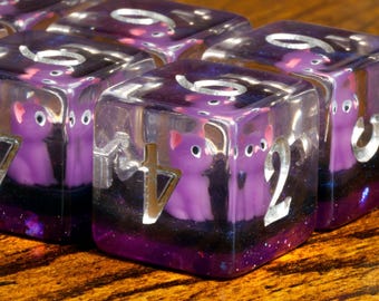 May include: A set of clear resin polyhedral dice featuring embedded miniature purple cat figurines. Each die has a dark purple and black glitter base with silver-colored numbers engraved on the faces. The dice are arranged in a cluster.