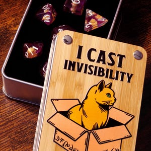 Könnte beinhalten: Eine Holzkiste mit einer Illustration einer Katze in einer Kiste und dem Text "I cast invisibility". Die Kiste hat einen Metallverschluss und ist mit einem Bambusmuster verziert. Die Kiste ist geöffnet und enthält einen Satz violetter und goldener Würfel.