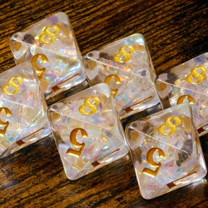 May include: Six clear, square dice with gold-coloured numbers and symbols. The dice have iridescent, colourful inclusions. The number 5 and the symbol 8 are visible on the dice. The dice are arranged on a dark, wooden surface.