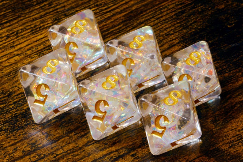 May include: Six clear, square dice with gold-coloured numbers and symbols. The dice have iridescent, colourful inclusions. The number 5 and the symbol 8 are visible on the dice. The dice are arranged on a dark, wooden surface.