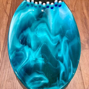 May include: An oval decorative dish in turquoise and teal. The dish has a swirling, marbled pattern, a dark grey border, and small white and blue accents along the top edge. It sits on a wooden surface. The dish's dimensions are approximately 25 cm in length.