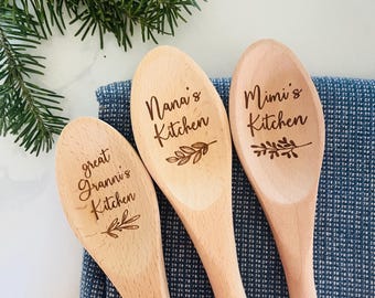 May include: Three wooden spoons with engraved text: "great granni's Kitchen", "Nana's Kitchen", and "Mimi's Kitchen". Each spoon has a decorative leaf design. The spoons are arranged on a blue patterned cloth, with greenery in the background.
