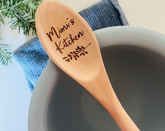May include: A wooden spoon with the text "Mimi's Kitchen" and a decorative sprig. The spoon is light brown and rests on a gray bowl with a blue cloth and a sprig of greenery in the background. Kitchenware.
