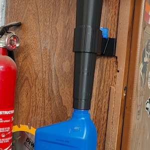 May include: A red funnel with a black extension attached to a blue plastic bottle. The funnel is mounted on a wall with a black bracket.