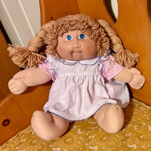 May include: A soft-bodied doll with light skin, bright blue eyes, and dimpled cheeks. The doll has yarn hair styled in two braided pigtails and wears a white dress with a blue scalloped collar, short pink and white striped sleeves, and a button.