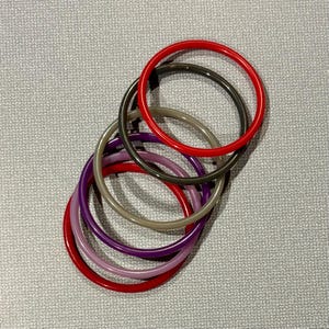 May include: A collection of seven colorful bangles in shades of red, purple, pink, gray, and black. The bracelets are circular and appear to be made of a smooth, glossy material. They are arranged in a slightly overlapping pattern against a textured gray background.