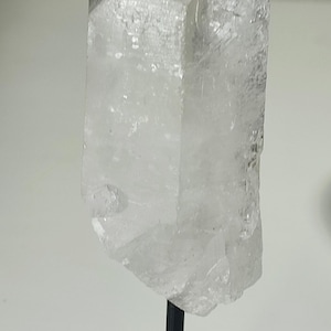 Könnte beinhalten: Eine klare Quarzkristallspitze auf einem schwarzen Metallständer. Der Kristall ist klar und hat eine leicht trübe Ausstrahlung. Der Ständer ist ein schlichtes Design mit einem quadratischen Sockel.