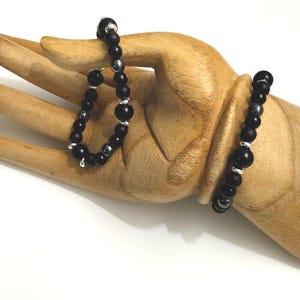 Puede incluir: Dos pulseras de cuentas, una negra con cuentas plateadas y otra negra con cuentas plateadas y un dije plateado. Las pulseras est&aacute;n en una mano de madera.