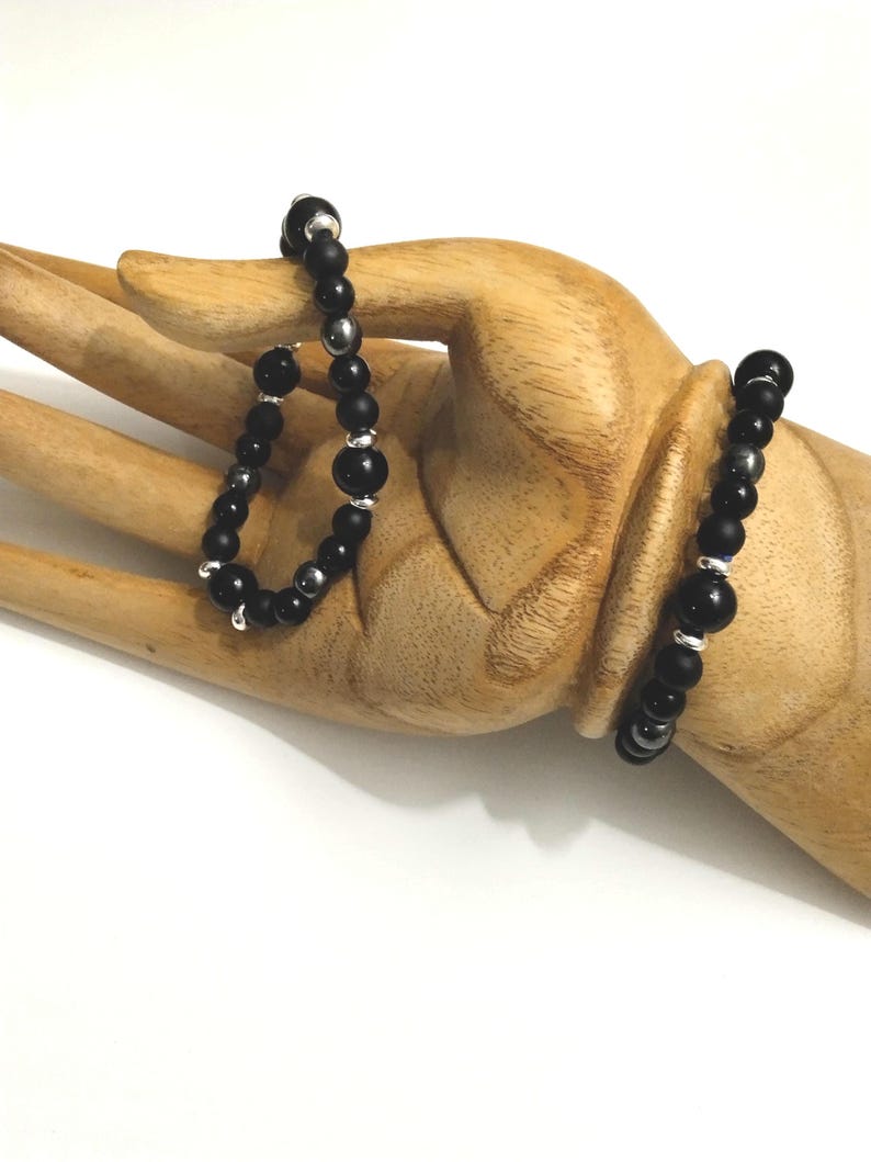 Puede incluir: Dos pulseras de cuentas, una negra con cuentas plateadas y otra negra con cuentas plateadas y un dije plateado. Las pulseras est&aacute;n en una mano de madera.