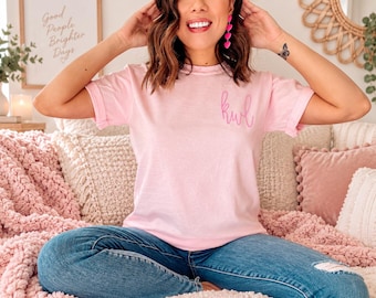 Puede incluir: Una camiseta de manga corta de color rosa claro con un pequeño monograma en cursiva de un tono rosa más oscuro en el lado izquierdo del pecho. La camiseta tiene cuello redondo y se combina con unos vaqueros azules con un detalle desgastado.