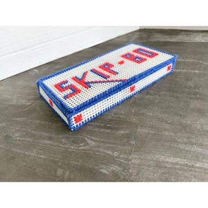 May include: A rectangular plastic canvas box with a lid, featuring a white grid base stitched with blue and red yarn. The top of the lid displays the text SKIP-BO in red and blue stitched lettering, with small red star accents on the sides.