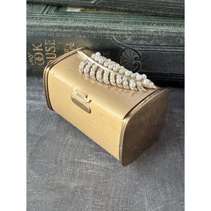 May include: A rectangular gold-toned metal box with a hinged lid. The top of the lid features a decorative gold-colored metal feather ornament embellished with numerous small white faux pearls arranged in a row along the center.