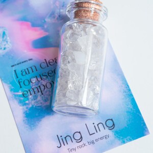 May include: A small glass bottle filled with clear quartz crystals. The bottle has a cork stopper and is sitting on a blue and pink background with the text "I am clear focused empowered" and "Jing Ling Tiny rock, big energy."