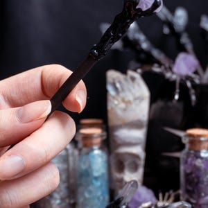 Puede incluir: Una varita negra con un cristal morado en la parte superior. La varita se sostiene en una mano y hay otros cristales y botellas en el fondo.