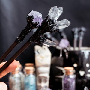 Puede incluir: Un par de palillos negros con detalles de cristal y amatista. Los palillos se sostienen en una mano y est&aacute;n enfocados. El fondo est&aacute; desenfocado y muestra varias botellas peque&ntilde;as de vidrio llenas de cristales.