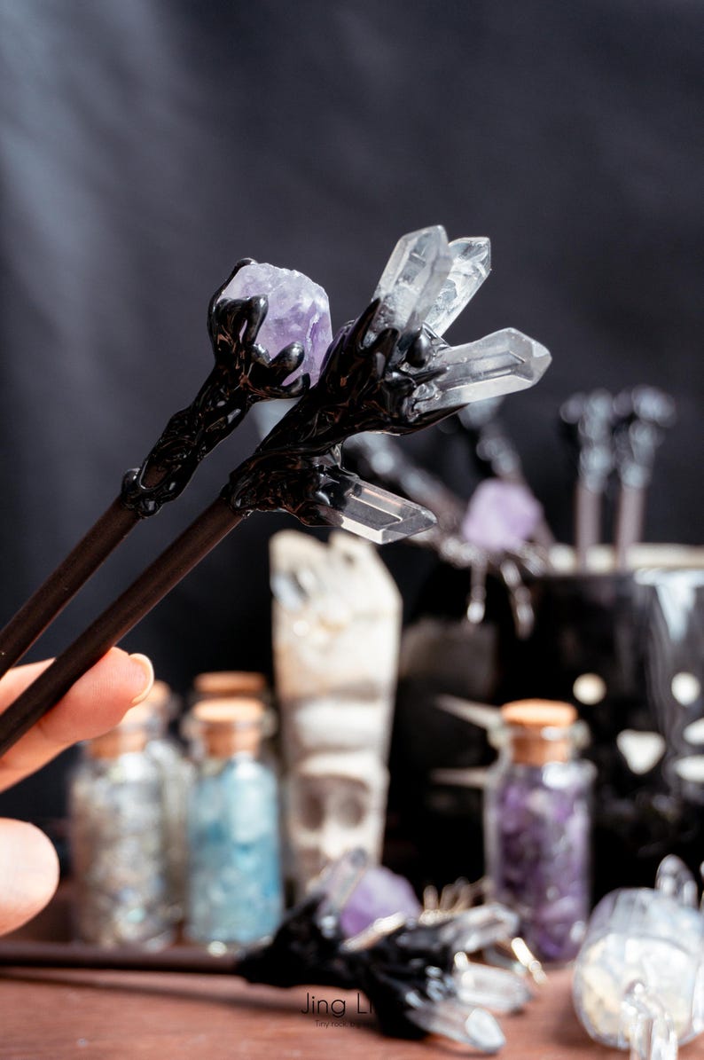 Puede incluir: Un par de palillos negros con detalles de cristal y amatista. Los palillos se sostienen en una mano y est&aacute;n enfocados. El fondo est&aacute; desenfocado y muestra varias botellas peque&ntilde;as de vidrio llenas de cristales.