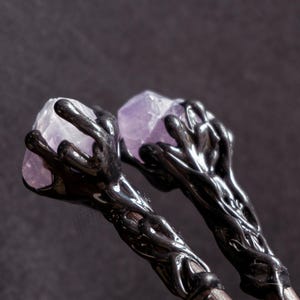 Puede incluir: Dos varitas negras con dise&ntilde;os intrincados, cada una coronada con un cristal de amatista p&uacute;rpura.