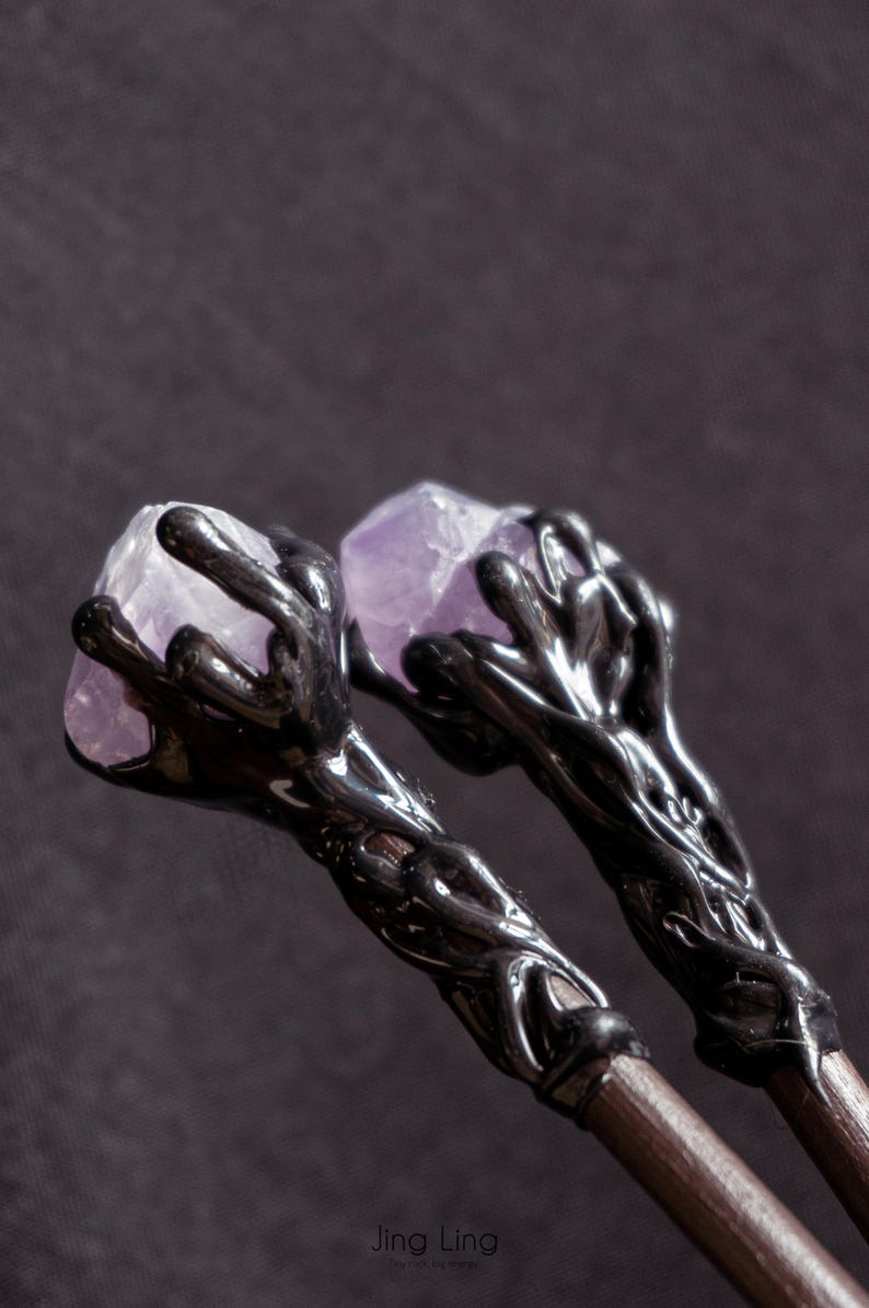 Puede incluir: Dos varitas negras con dise&ntilde;os intrincados, cada una coronada con un cristal de amatista p&uacute;rpura.