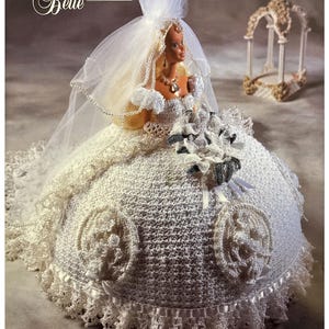 May include: A white crocheted bridal gown with a lace trim and a white veil. The gown is designed for a Barbie doll and features a floral design. The doll is holding a bouquet of white flowers. The text "The Bridal Belle Collection" is visible in the image.