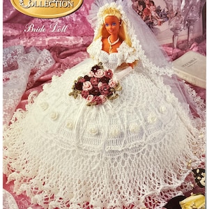 May include: A white crocheted bridal gown for a doll, with a white veil and a bouquet of pink and white roses. The doll is wearing a white dress with a pearl necklace and a cross pendant. The doll has blonde hair and blue eyes. The image is from the "Gems of the South Collection" and is titled "Bride Doll".